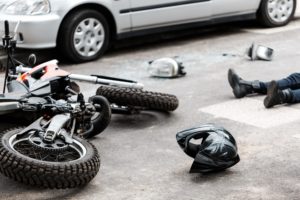motorcycle crash scene with motorcycle, motorcyclist boot and helmet laying on road in front of a white parked car