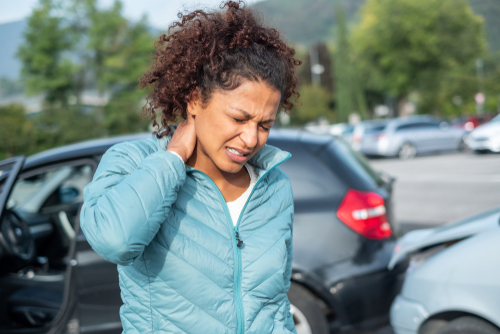 A young person is talking on the phone while outdoors. They are next to a family car parked on the street, surrounded by trees. The individual is wearing a jacket and appears engaged in conversation.