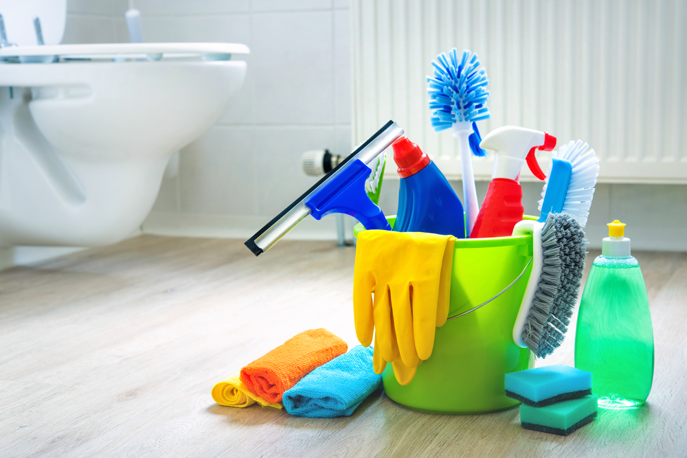 A green bucket filled with cleaning supplies sits on a bathroom floor next to a toilet.