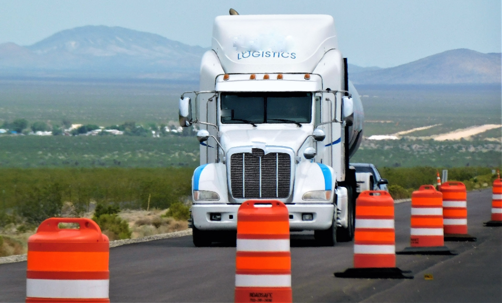 The image shows a truck navigating around an orange and white traffic cone, likely on a road or highway, with tags related to freight transport, commercial vehicles, and trailer trucks.