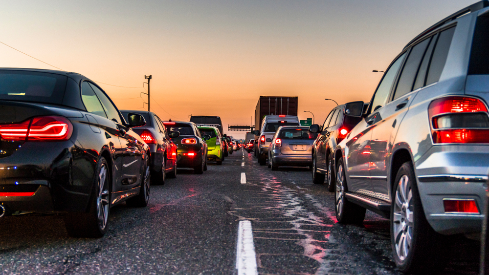 A line of cars of various makes and models are stopped in a traffic queue on a road. The cars are dark-colored, including black, silver, and dark gray.