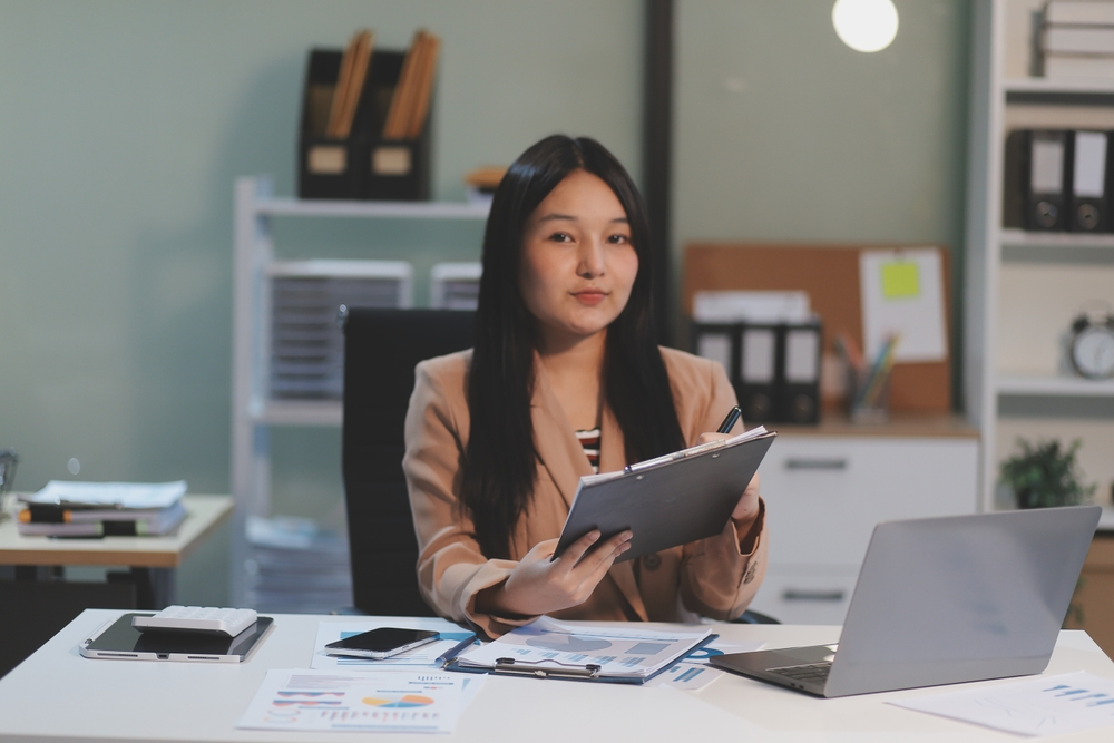 A woman dressed in a business suit is seated at a desk, engaged with her laptop, representing professionalism and productivity.