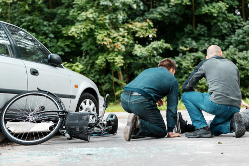 Two people assist an injured individual who was hurt in a collision between a bicycle and a car.