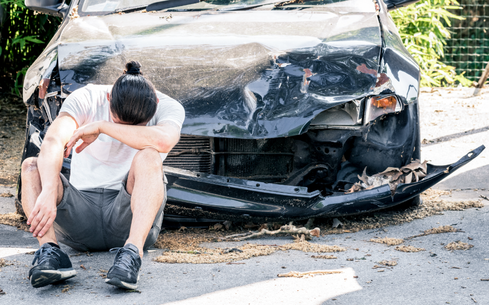 A man sits on the ground beside a damaged car, reflecting on the incident that caused the vehicle's condition.