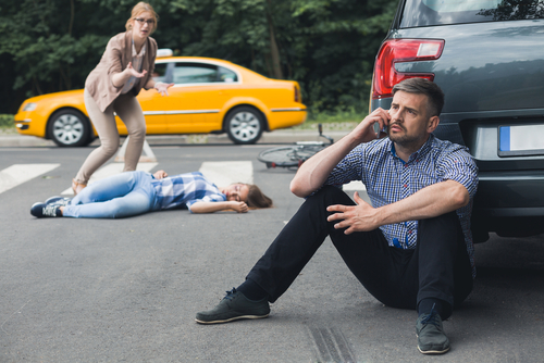 A man sits on the ground next to a car, observing a woman lying on the ground, indicating a serious or urgent situation.